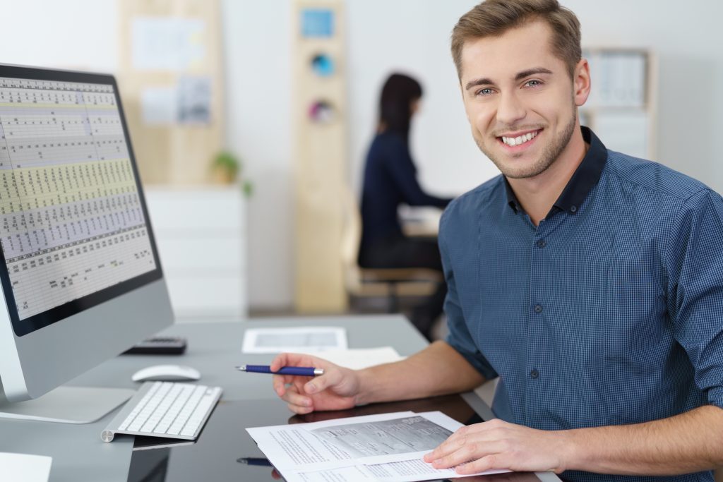 Happy successful young businessman at his desk in the office working on a report looking at the camera with a warm smile